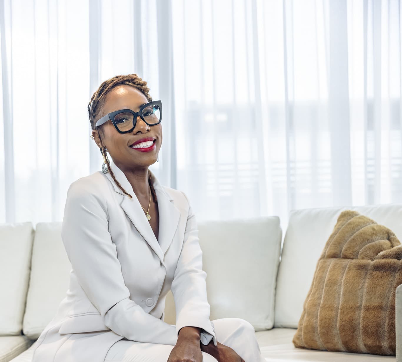 Dr. Natalie Edwards-Hudson, wearing a cream blazer suit and glasses, smiling while seated on a couch in front of sheer curtains